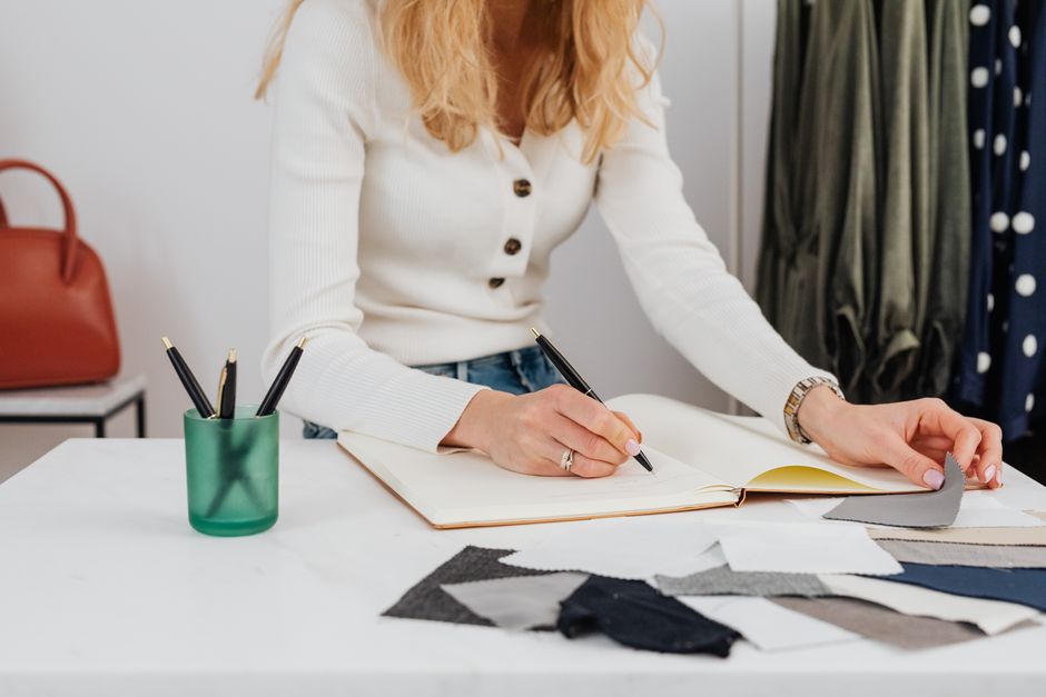 A woman designer sketches fashion ideas on a sketchpad with fabric swatches nearby.